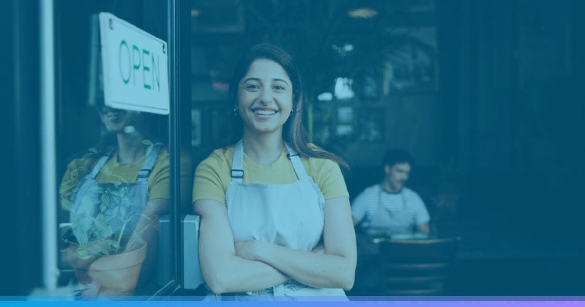 Smiling woman in apron stands at cafe entrance with an open sign on the door.