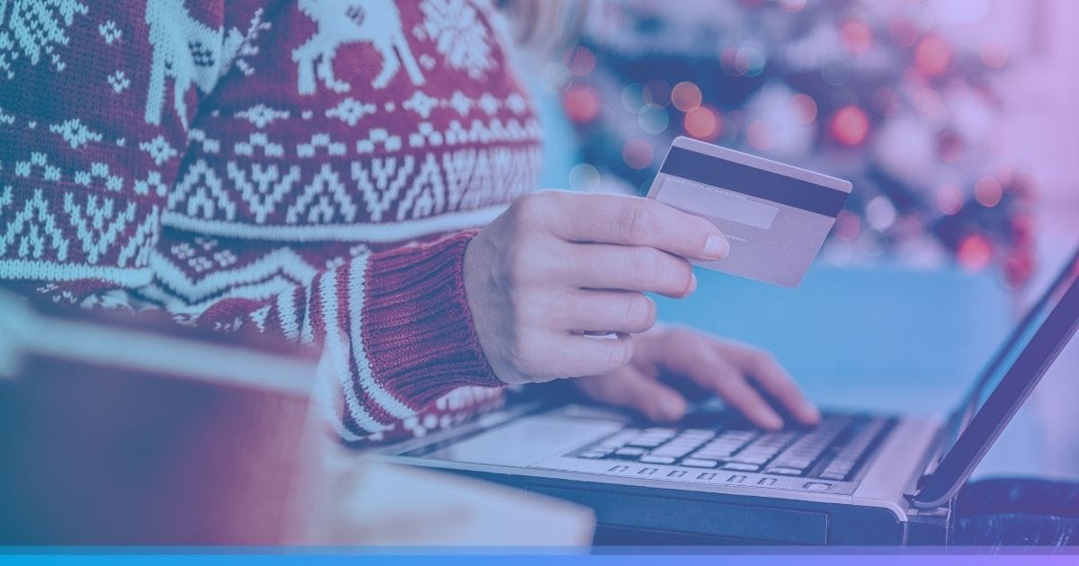 Person wearing a festive sweater shopping online with a credit card on a laptop, holiday decorations in the background.