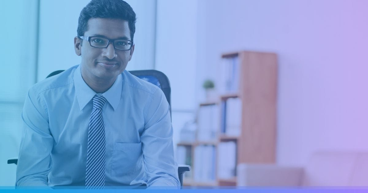 Man in blue shirt and glasses sitting in office chair, smiling, with blurred shelves in background.