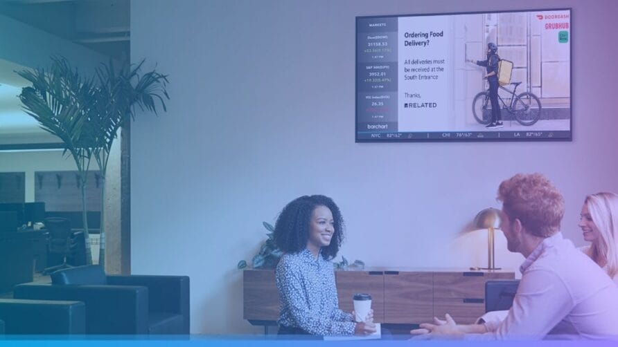 People sitting in a modern office lounge discussing with a digital sign displaying food delivery information on the wall.