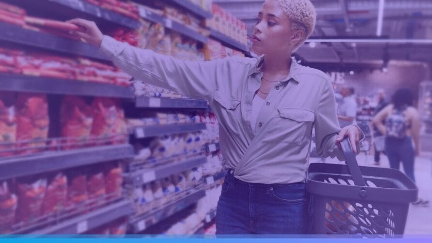 Person shopping in a supermarket aisle, reaching for a product on the shelf, holding a basket.