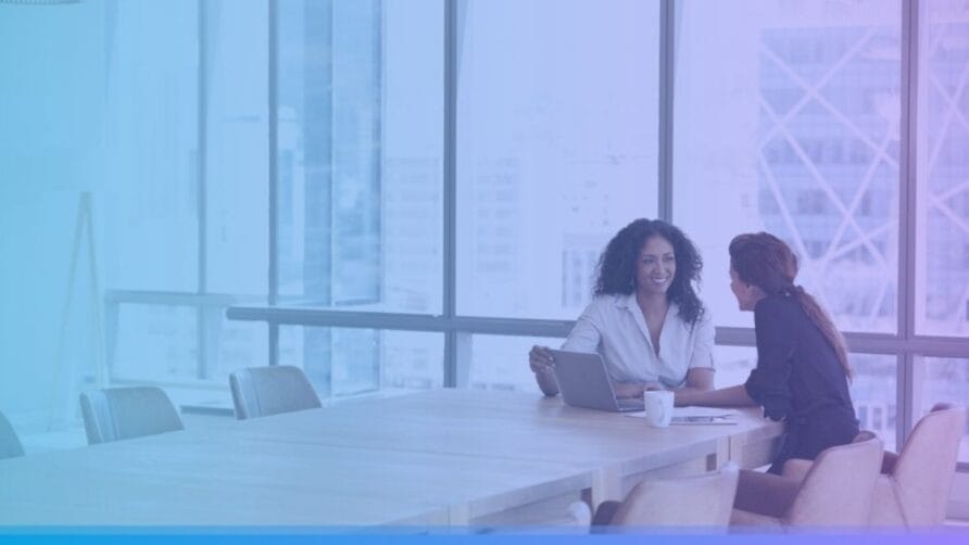 Two women sitting at a conference table in an office, discussing work with a laptop open in front of them.