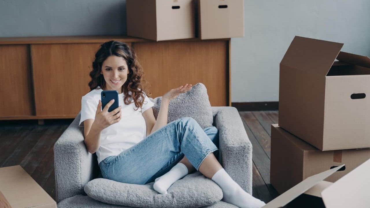Woman sitting in armchair using smartphone, surrounded by unpacked cardboard boxes in a new home setting.