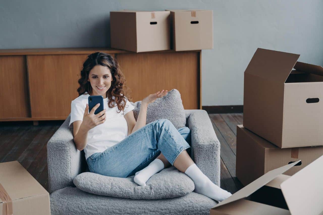 Woman sitting in armchair using smartphone, surrounded by unpacked cardboard boxes in a new home setting.