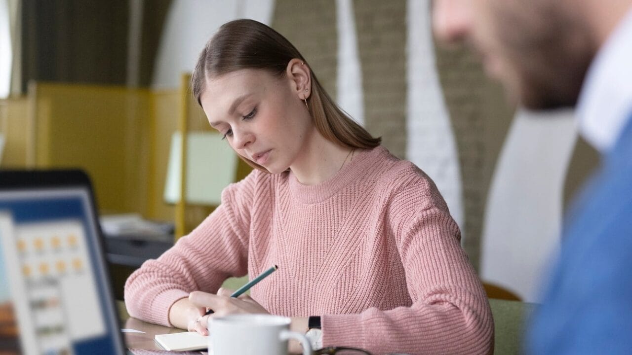 Woman writing notes at desk with coffee, while a man types on a laptop in the foreground.
