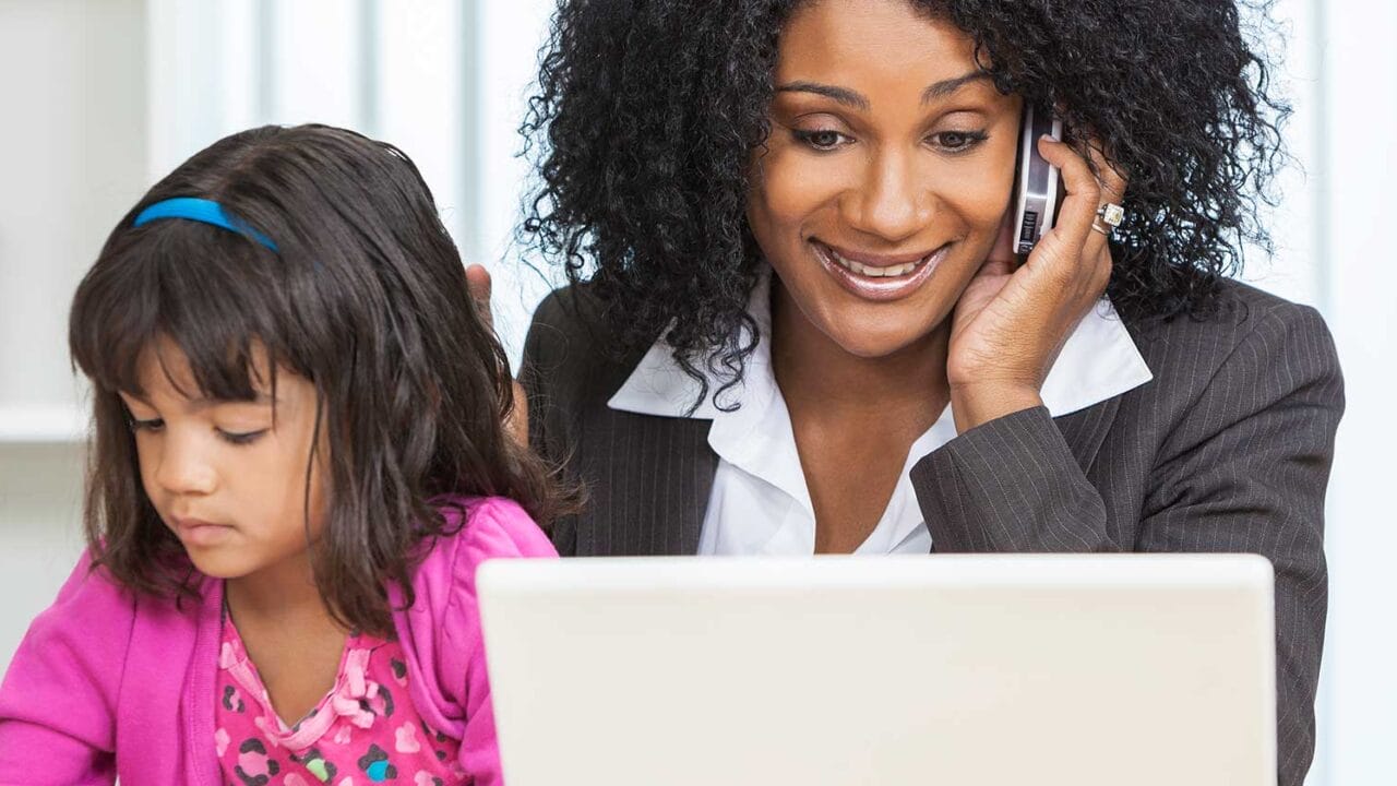 Woman working on a laptop and talking on the phone while a young girl draws next to her, highlighting multitasking.