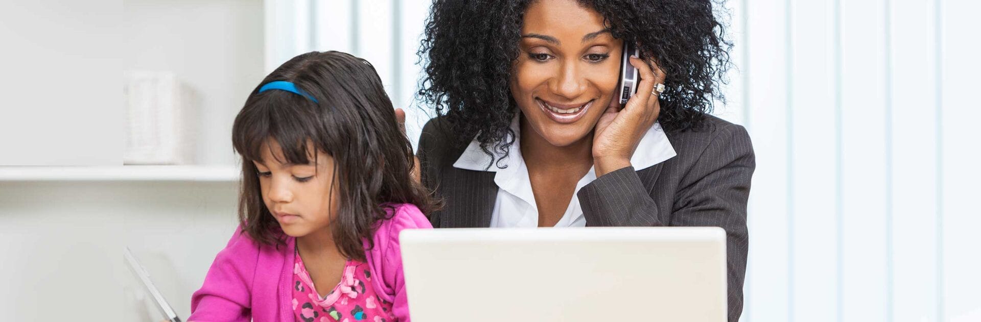 Woman working on a laptop and talking on the phone while a young girl draws next to her, highlighting multitasking.