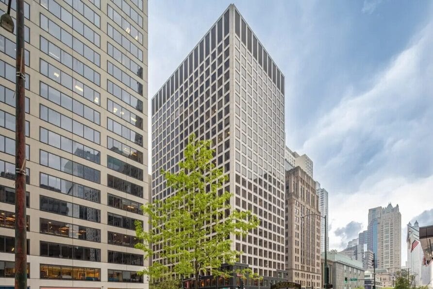 Tall modern office building with a grid-like window pattern, surrounded by other buildings and a small leafy tree in the foreground.
