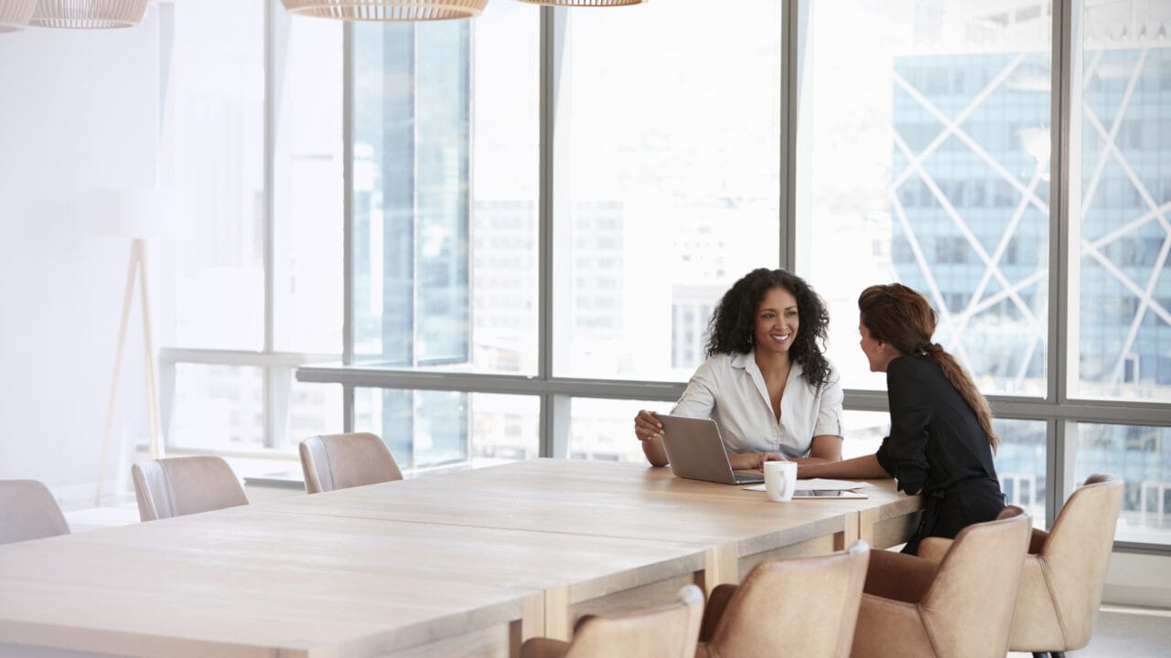 Two women sitting at a long table in a modern office, engaged in discussion with a laptop open in front of them.