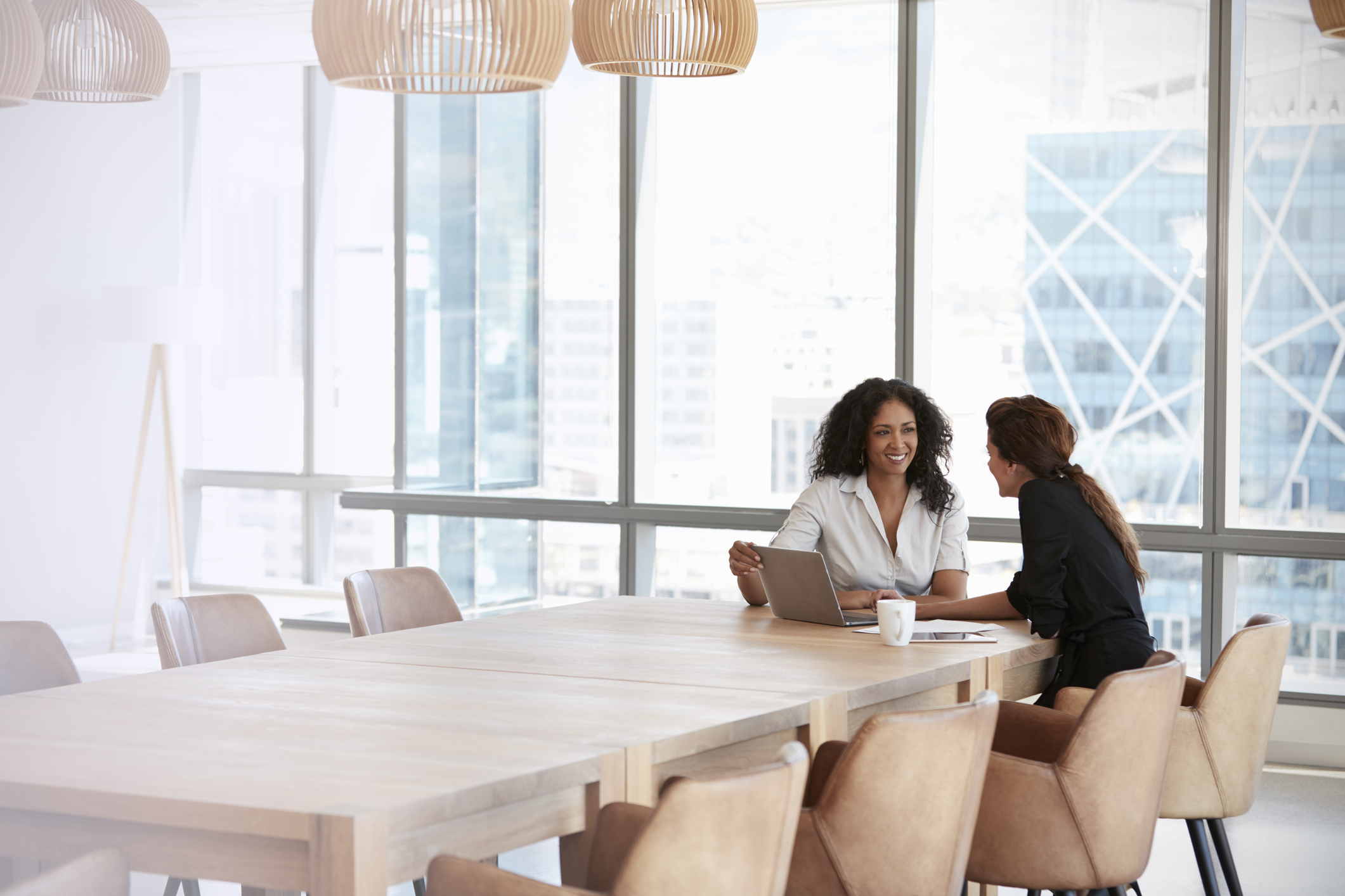 Two women sitting at a long table in a modern office, engaged in discussion with a laptop open in front of them.