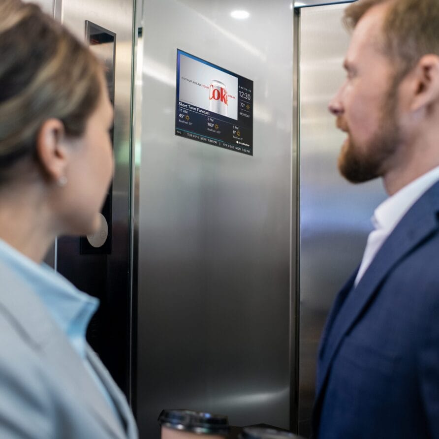 Two people in business attire standing in an elevator, looking at a digital screen displaying information.