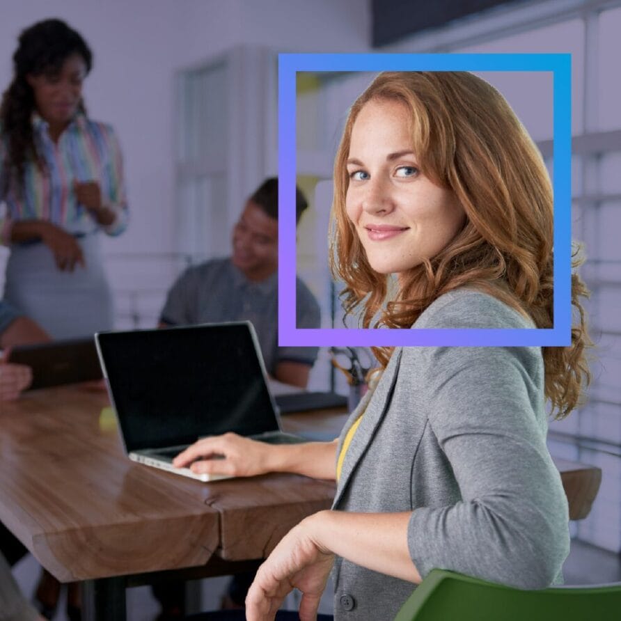 Woman smiling at the camera in a meeting room with a laptop, colleagues in the background.