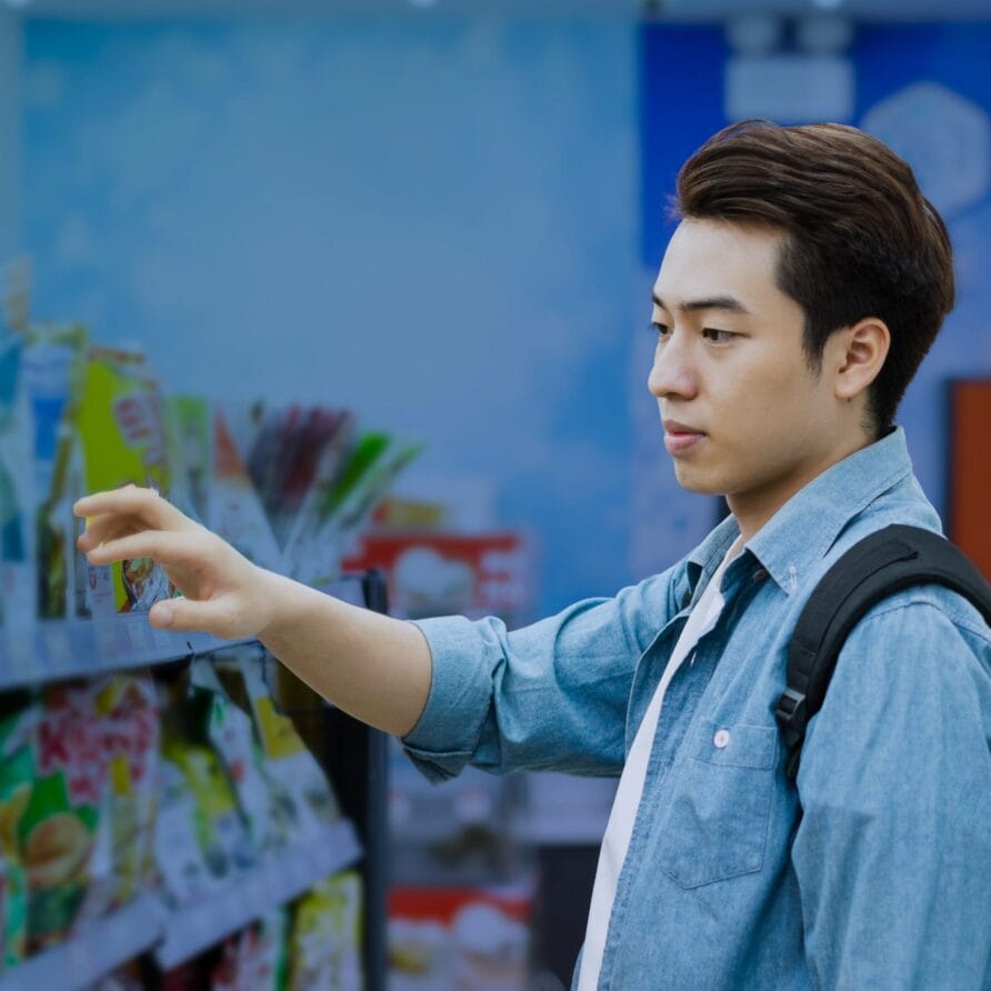 Person with a backpack shopping in a store aisle, reaching for items on a shelf, wearing a blue shirt and focused expression.
