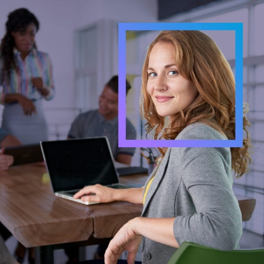 Woman in a meeting room with a laptop, highlighted by a digital frame overlay, colleagues talking in the background.