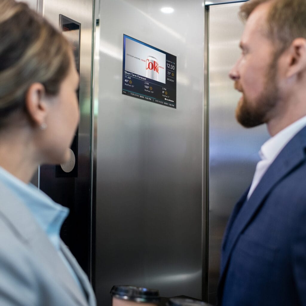 Man and woman standing in an elevator, looking at a screen displaying information.