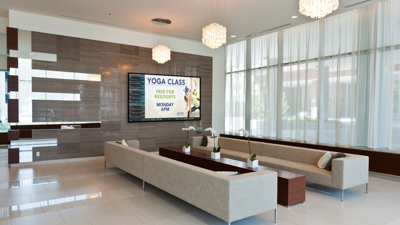 Modern lobby with beige sofas, elegant lighting, and a digital sign announcing a free yoga class for residents on Monday at 6 PM.