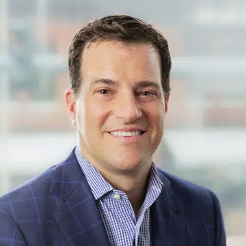 Man in a blue suit and checkered shirt smiling with a blurred office background.