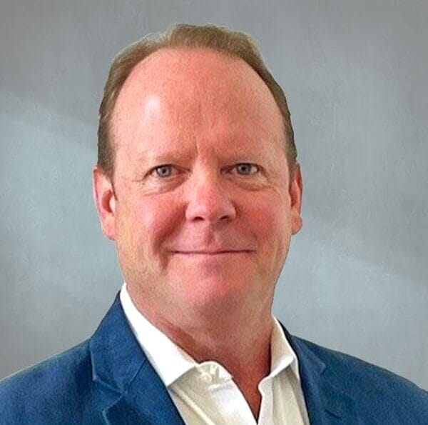 Man in a blue blazer with a light shirt standing against a gray background.