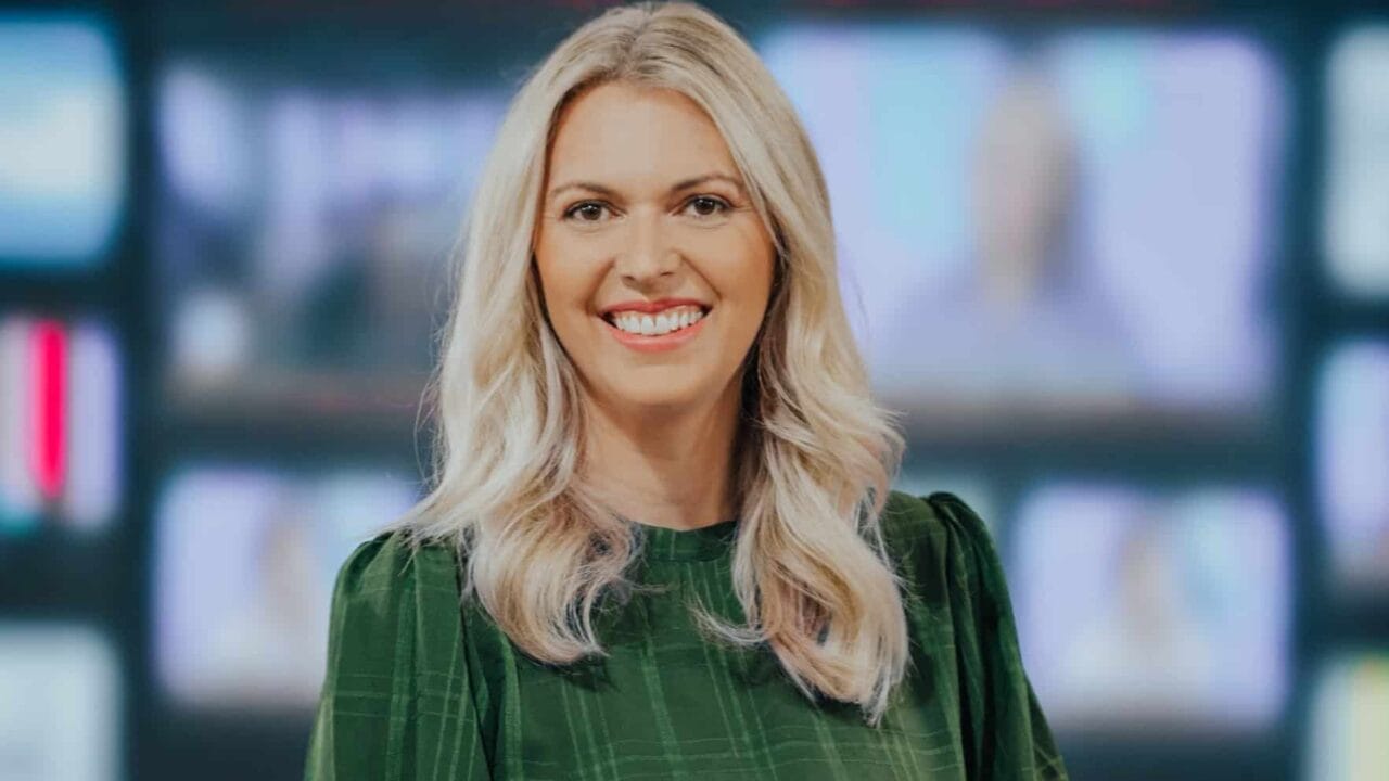 Smiling person with long blonde hair wearing a green checkered top in front of a blurred screen background.