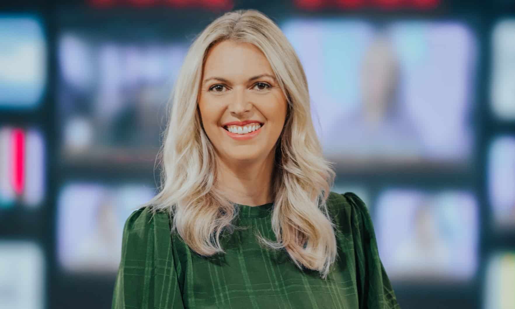 Smiling person with long blonde hair wearing a green checkered top in front of a blurred screen background.