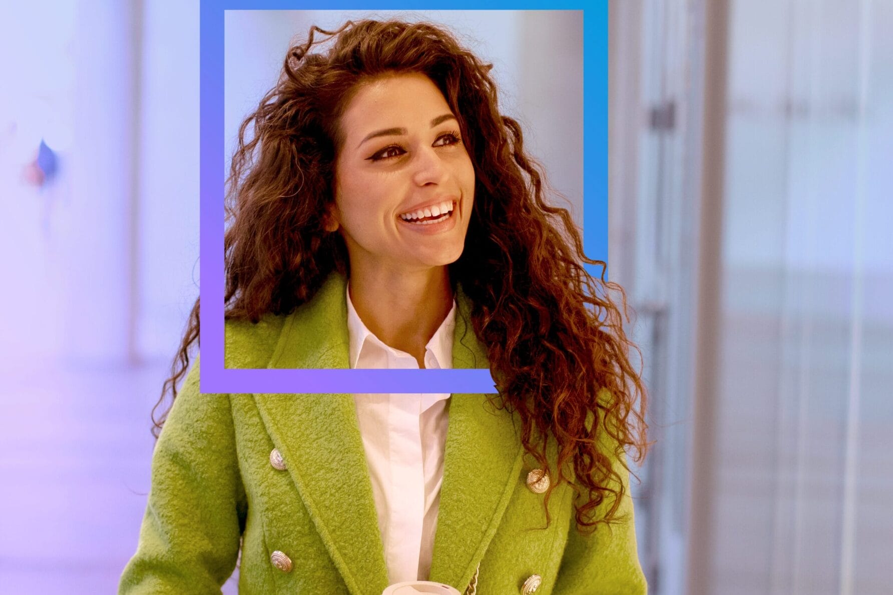 Woman with curly hair in a green coat holding a coffee cup, standing in a brightly lit hallway.