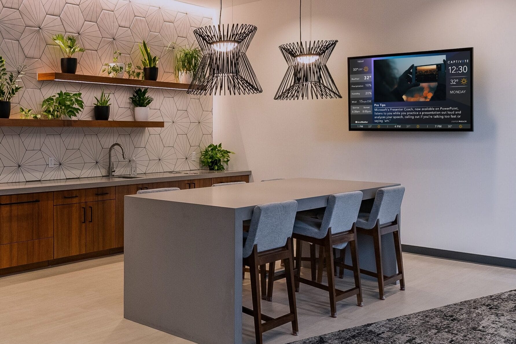 Modern kitchen with island, stools, wall-mounted TV, hanging lights, plants on shelves, and a patterned rug in neutral tones.