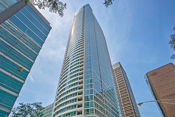 Tall modern skyscraper with curved glass facade under a clear blue sky, surrounded by other buildings and tree branches.