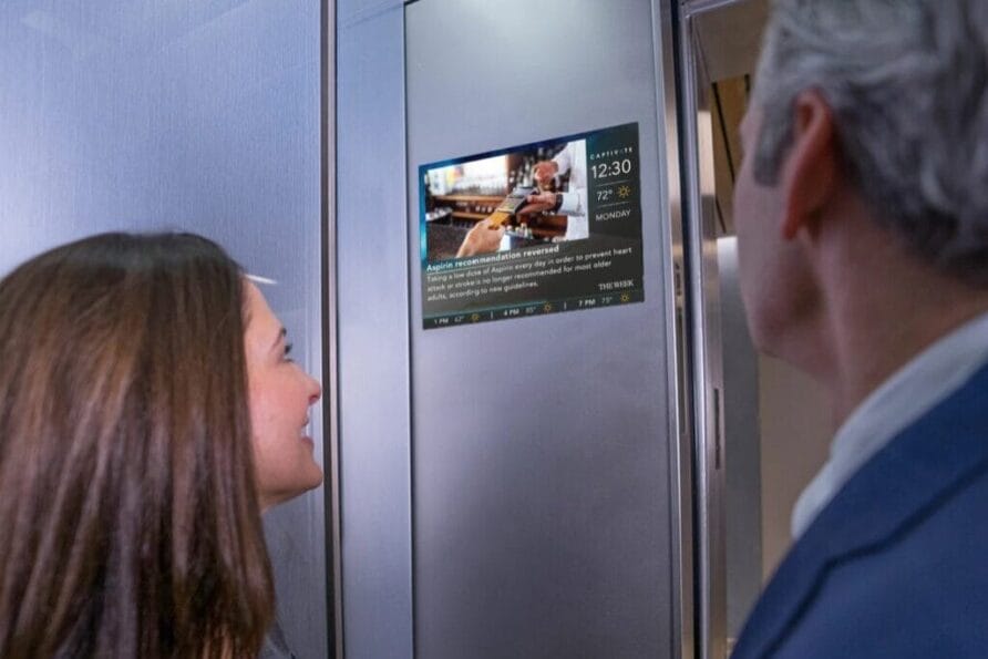 Two people looking at a digital screen in an elevator, displaying news and weather updates.