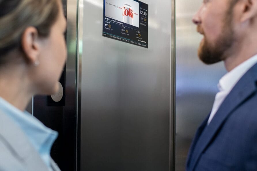 Two people in business attire standing inside an elevator looking at a screen displaying information.