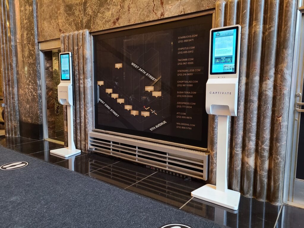 Two digital kiosks in a building lobby with a large directory map on the wall between them.