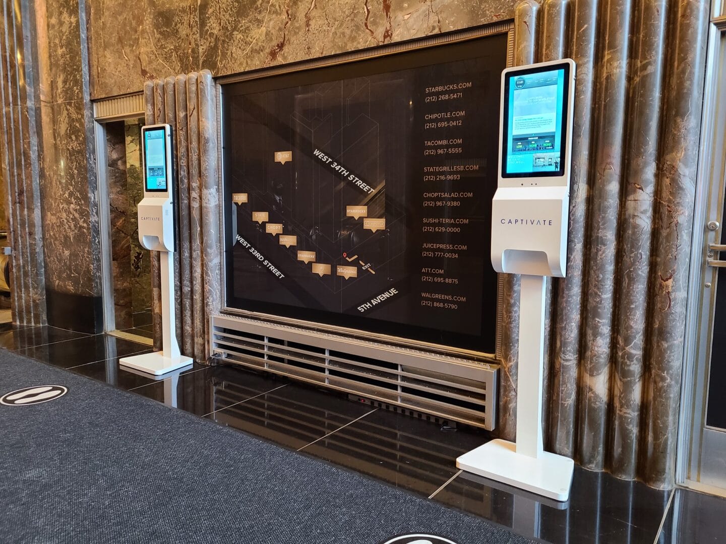 Two digital kiosks in a building lobby with a large directory map on the wall between them.