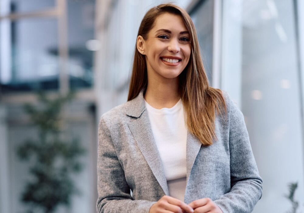 Smiling woman in a gray blazer standing outdoors near a modern glass building.