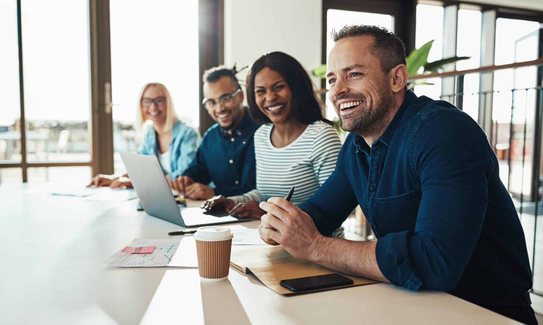 Four people smiling and working together at a table with laptops and coffee in a brightly lit office space.