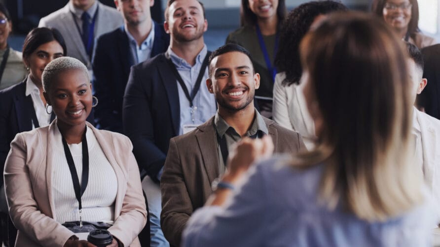 Diverse group of people smiling and listening to a speaker during a seminar or conference in a well-lit room.