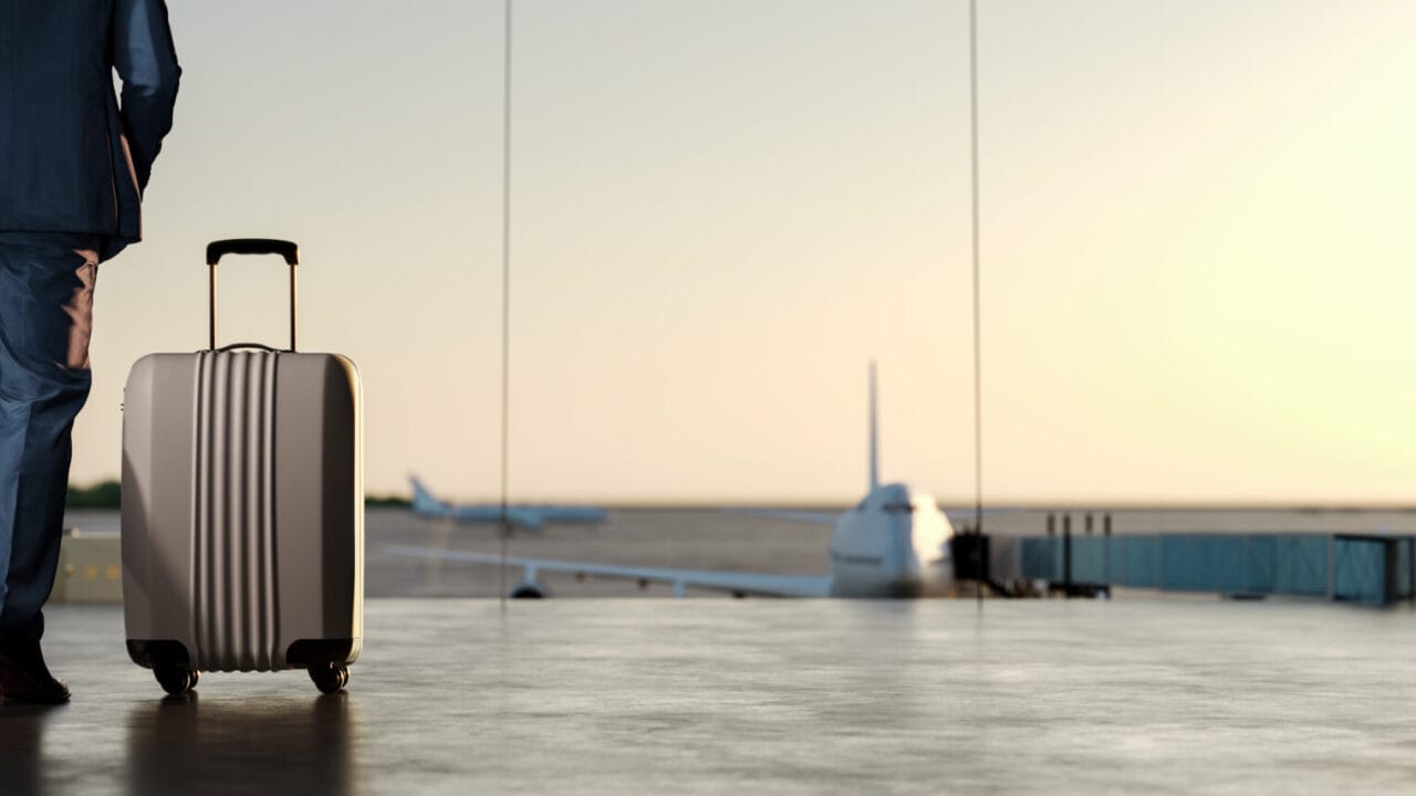 Traveler with suitcase at airport terminal, watching planes take off against a sunset sky.