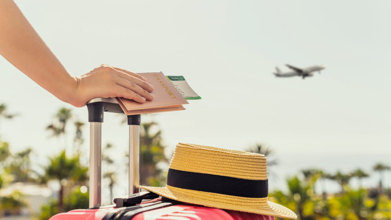 Hand holding a passport with tickets on pink suitcase; straw hat on suitcase; airplane flying in the background.