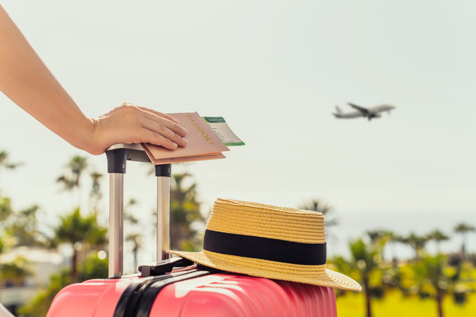 Hand holding a passport with tickets on pink suitcase; straw hat on suitcase; airplane flying in the background.