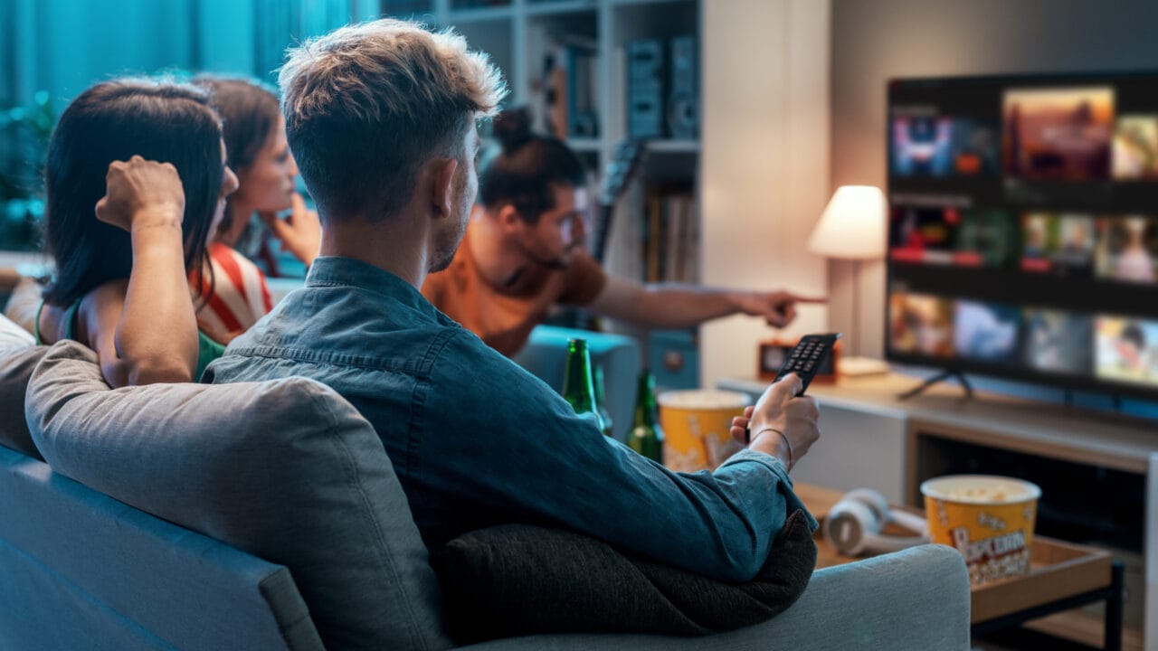 Four people sitting on a couch watching a show on TV with popcorn and drinks in a cozy living room setting.