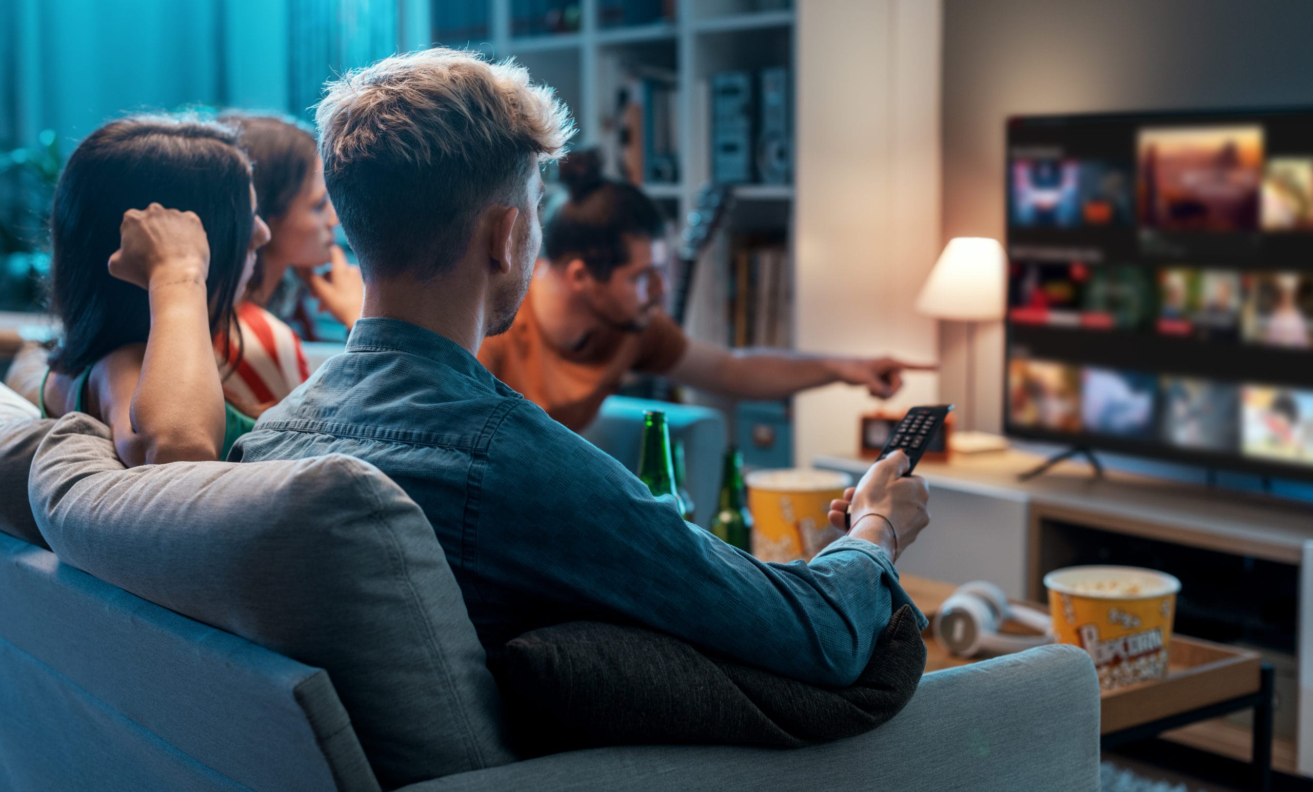 Four people sitting on a couch watching a show on TV with popcorn and drinks in a cozy living room setting.
