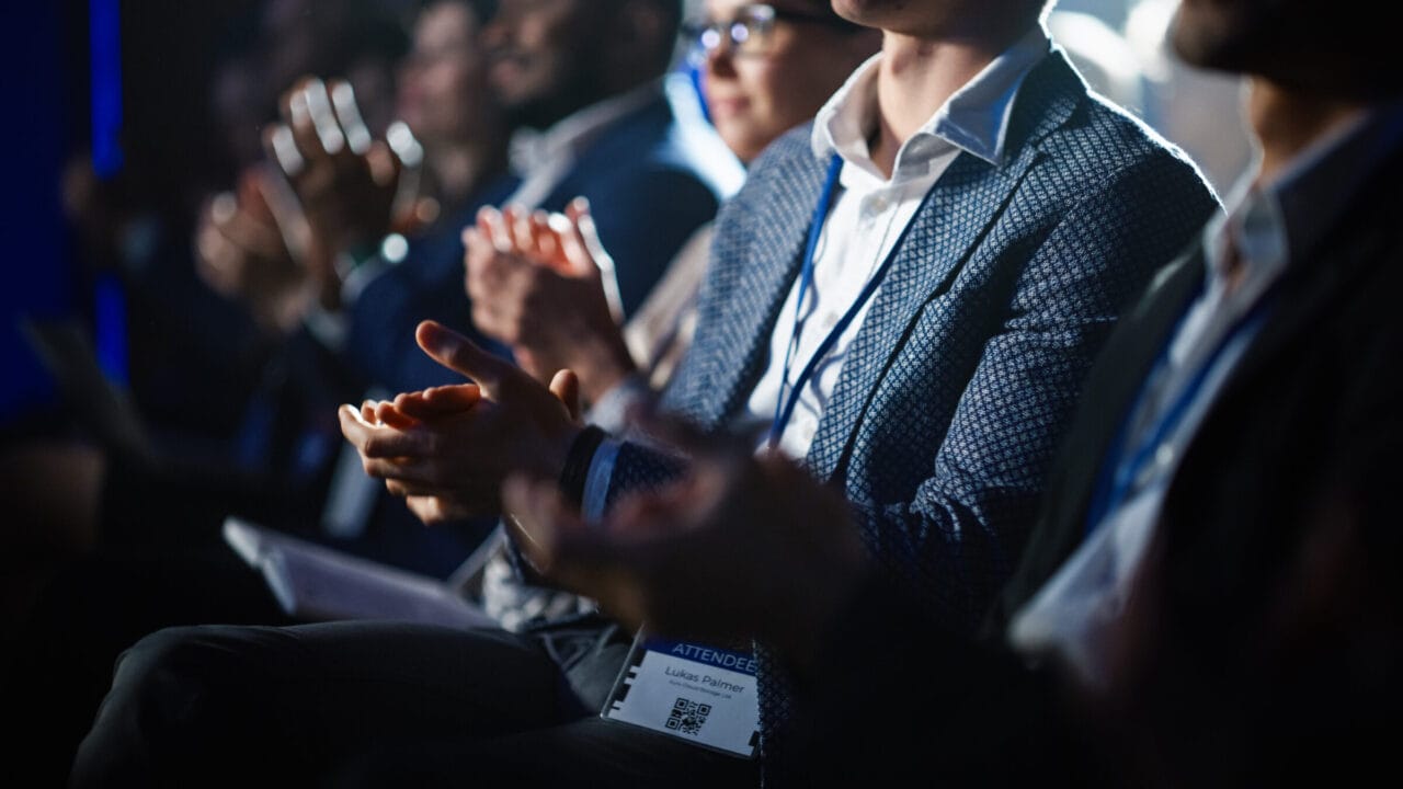 Audience members clapping at a conference or event, wearing business attire and name badges.