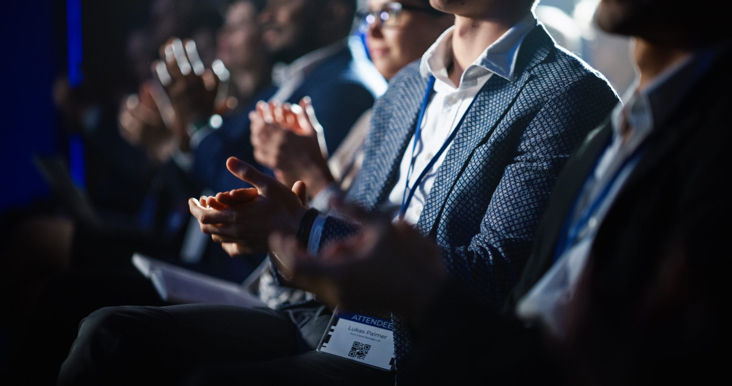 Audience members clapping at a conference or event, wearing business attire and name badges.