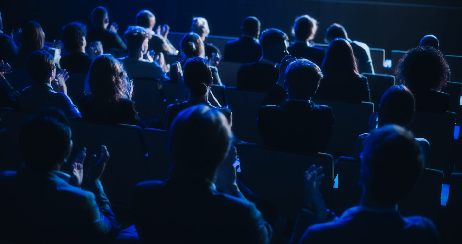Audience sitting in a dimly lit theater, clapping and watching a performance on stage with blue lighting.