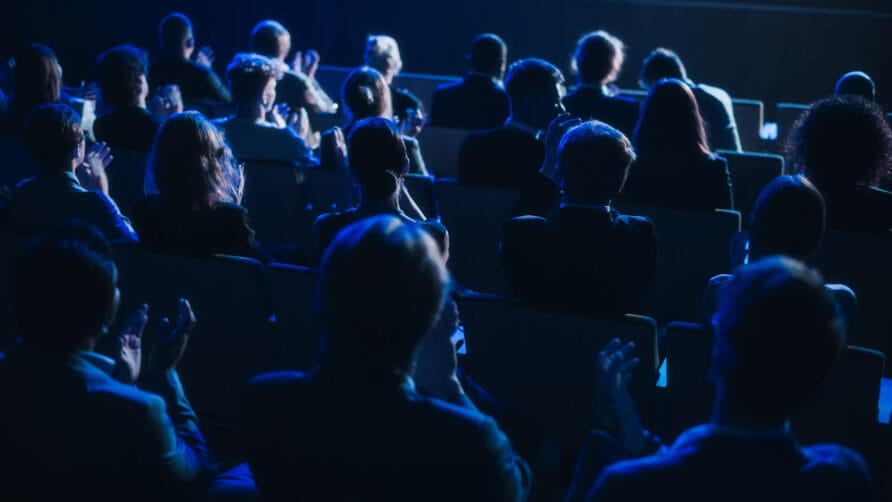 Audience sitting in a dimly lit theater, clapping and watching a performance on stage with blue lighting.