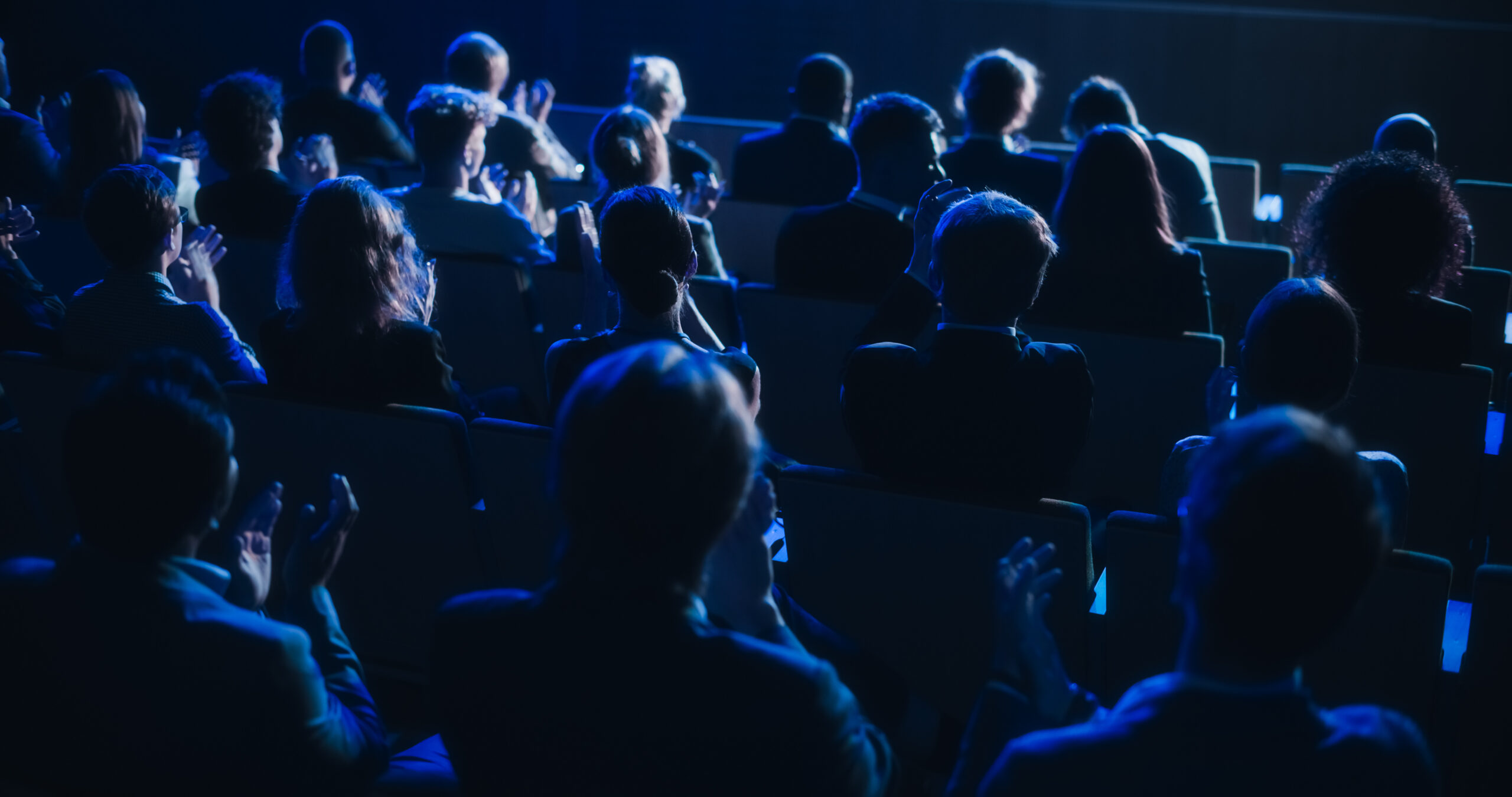 Audience sitting in a dimly lit theater, clapping and watching a performance on stage with blue lighting.