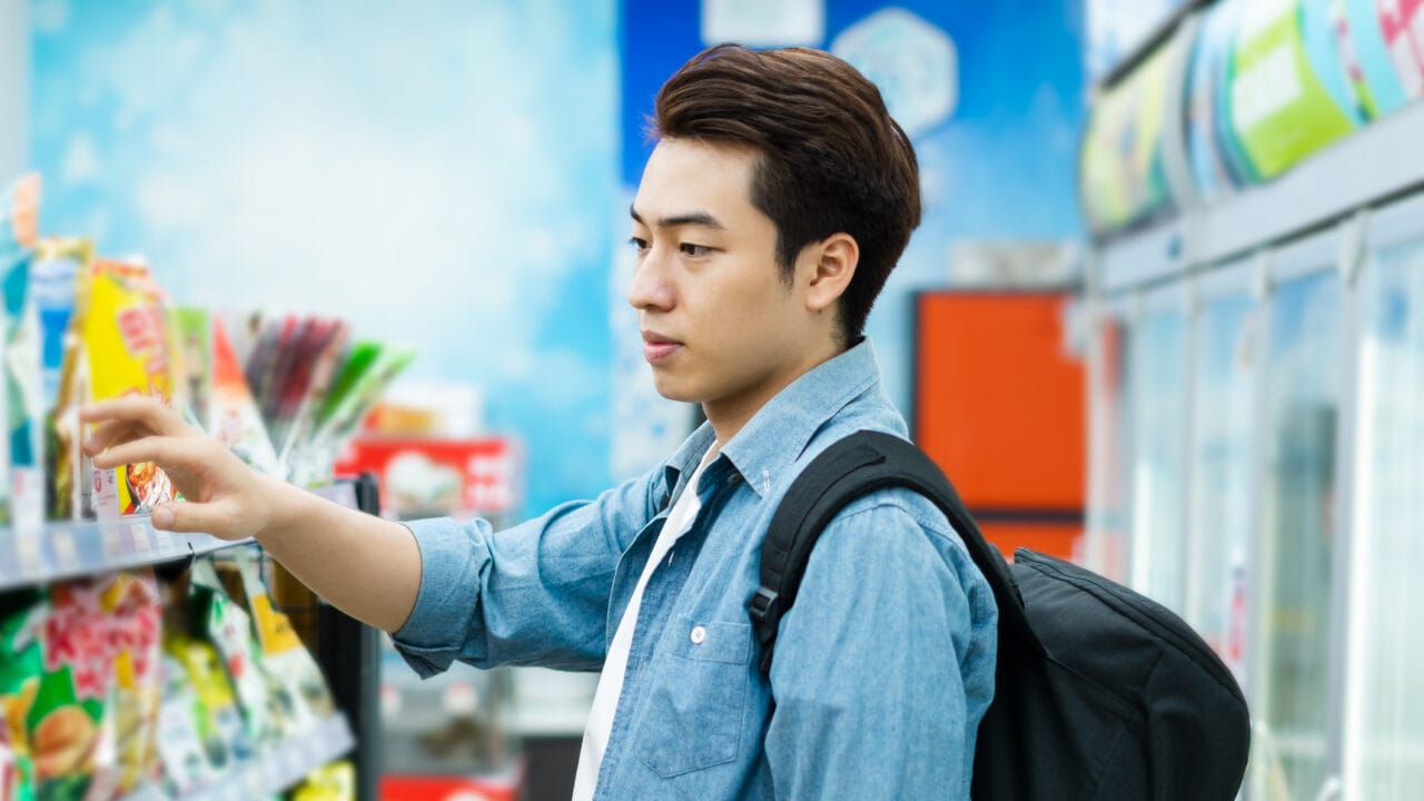 Person shopping for snacks in a convenience store, wearing a denim shirt and backpack, examining items on a shelf.