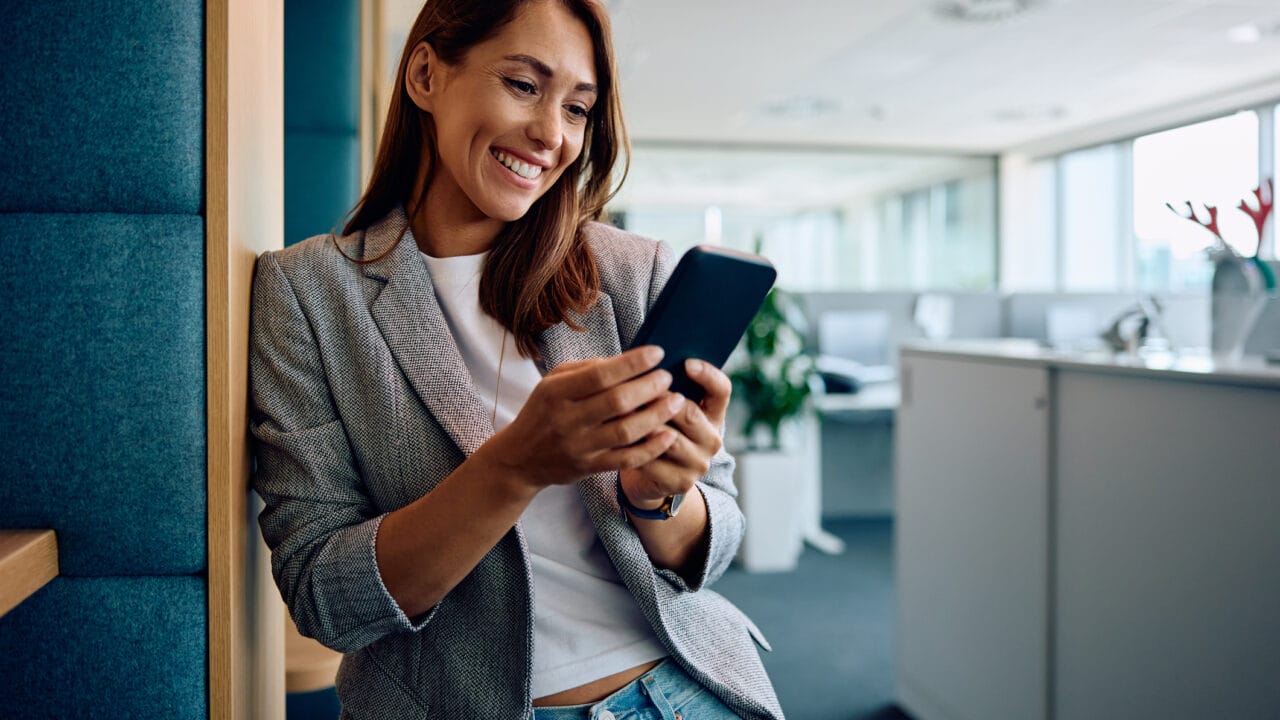 Woman smiling while using her smartphone in a modern office setting.