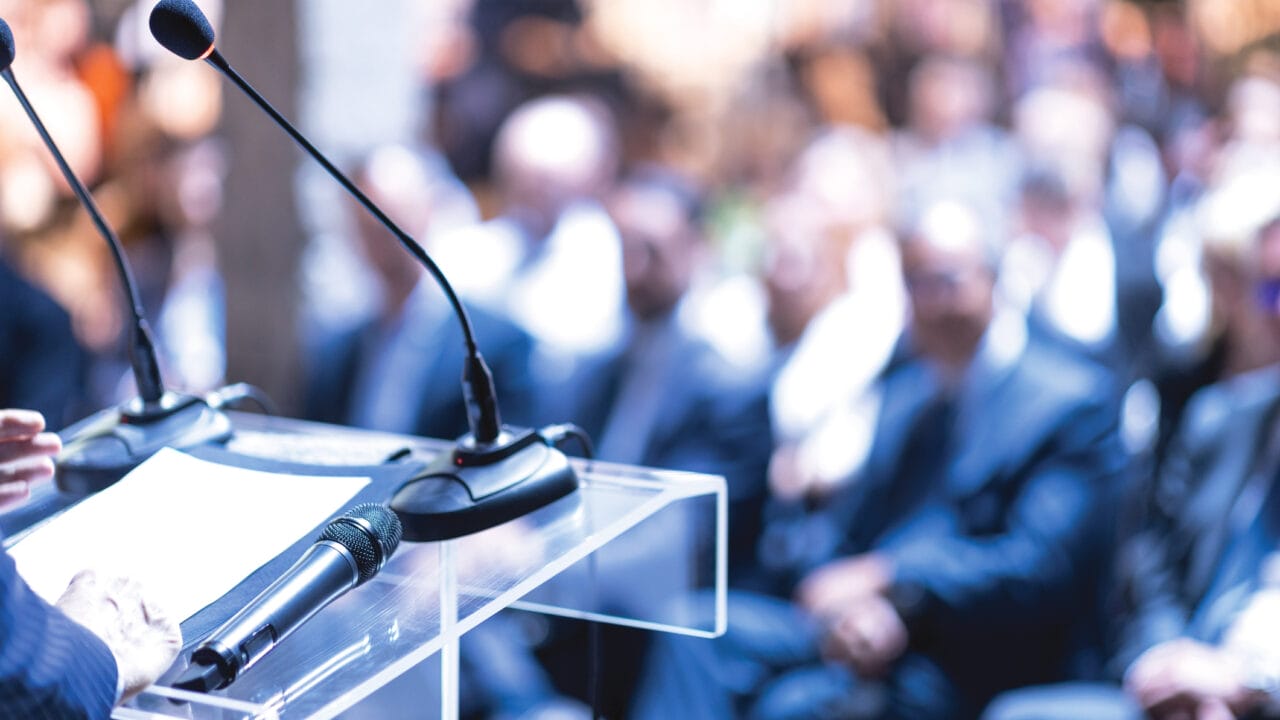 Speaker at a podium with microphones addressing a seated audience during a conference or event.