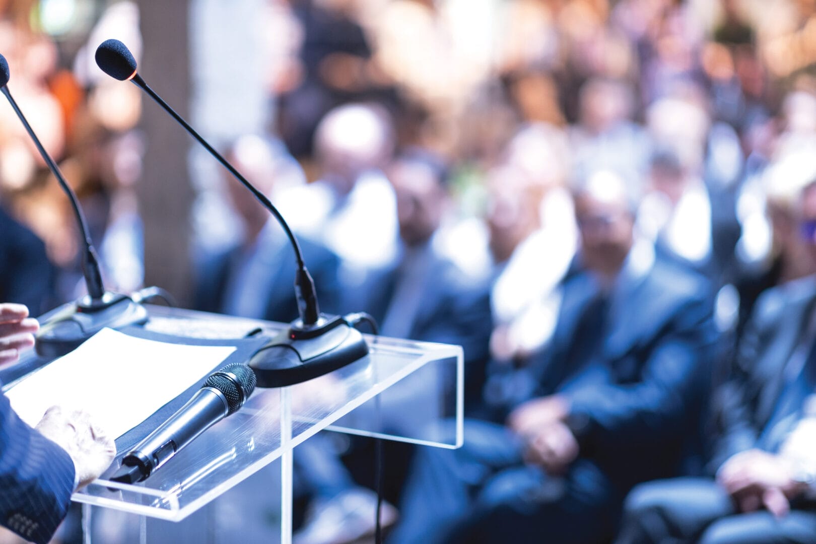 Speaker at a podium with microphones addressing a seated audience during a conference or event.