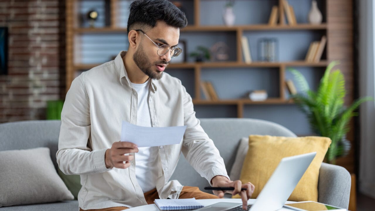 Man sitting on couch, wearing glasses, holding paper, working on a laptop with bookshelves and plants in the background.