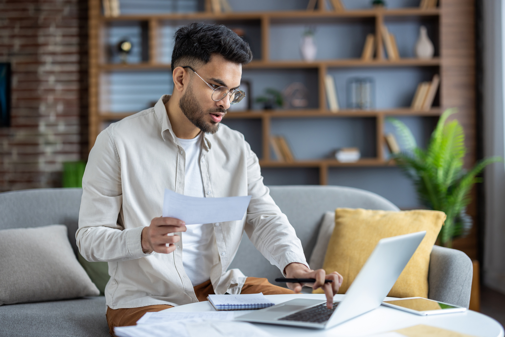 Man sitting on couch, wearing glasses, holding paper, working on a laptop with bookshelves and plants in the background.